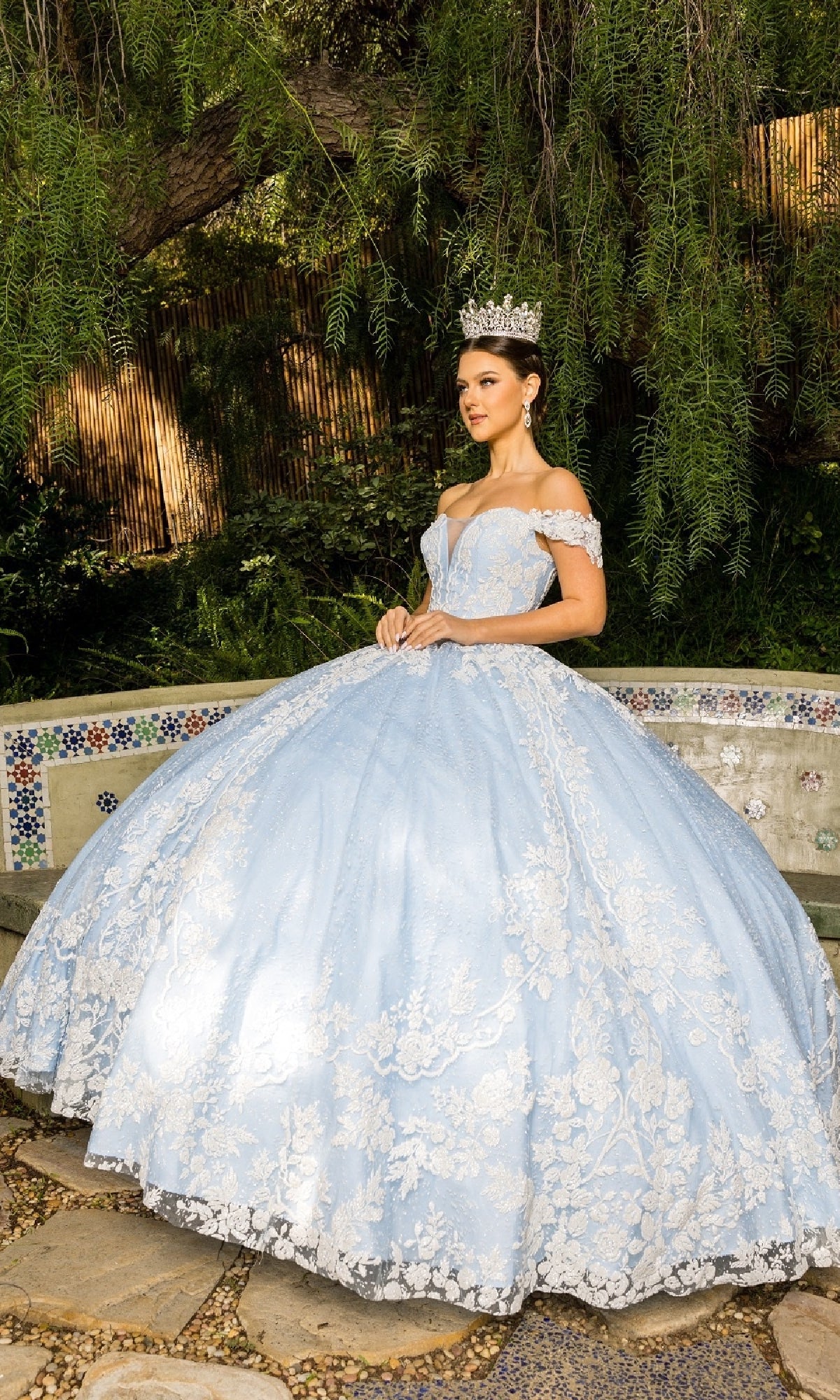 A young woman wearing the Cinderella Couture 8070J Quinceanera Dress, a light blue off-the-shoulder gown with floral lace embroidery, and a silver crown stands outdoors by greenery and a tiled bench.